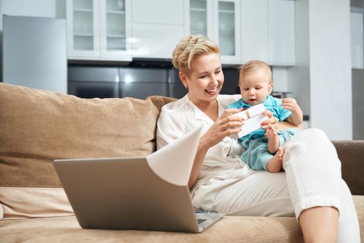 Cheerful mother holding cute baby on hands and opening gift box. Pretty woman sitting on couch with wireless laptop. Birthday celebration at home.
