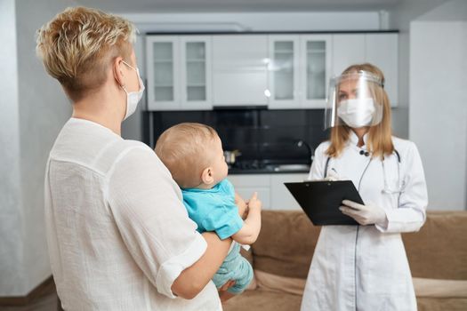 Back view of mother carrying baby on hands while pediatrician in medical uniform and mask doing examination. Health care and children concept.