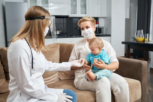 Competent doctor in protective mask and gloves visiting her little patient at home. Young mother sitting on couch with cute toddler i on hands. Medical treatment.