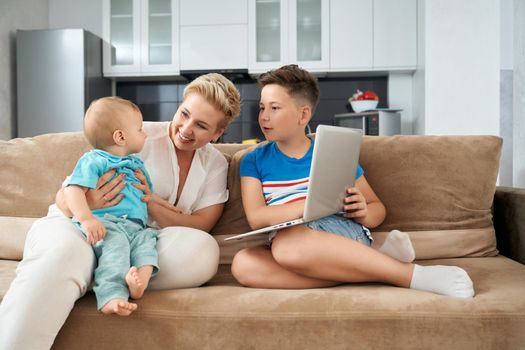 Smiling mother with two sons of different ages playing on couch and using modern laptop. Domestic lifestyle and family time.