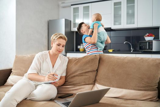 Pleasant woman with short blond hair sitting on couch and working on laptop. To happy boys playing and smiling on kitchen. Concept of household and work.