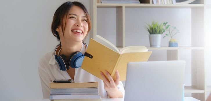 Portrait of smiling asian student girl and reading a book at homeschool before learning virtual internet online class. covid pandemic