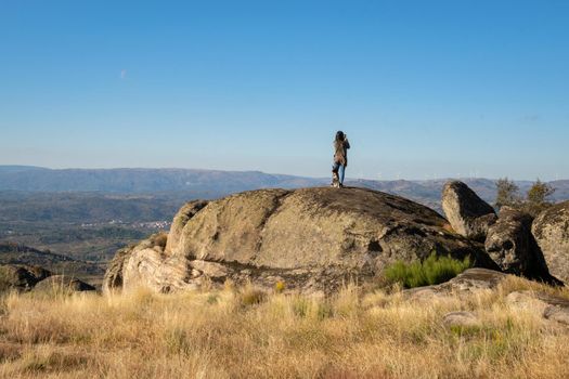 Caucasian young woman with brown dog on top of a boulder stone seeing Sortelha nature mountain landscape, in Portugal