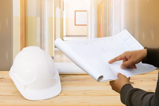 engineer finger point on paper plan blueprint in check building technician place construction site gypsum board wall interior background and white safety helmet plastic on wood floor table