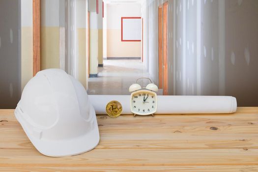 white safety helmet plastic, paper roll plan blueprint alarm clock time a rest at noon, bitcoin coin on wood floor table and gypsum board wall interior construction site background. with copy space add text