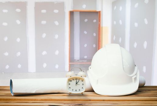 white safety helmet plastic, paper roll plan blueprint alarm clock time a rest at noon on wood floor table and gypsum board wall remodel interior with copy space add text