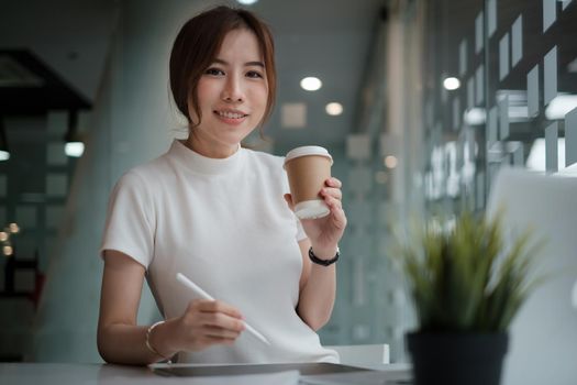 Cretive woman in white dress smiling holding stylus pencil and coffee in home office. Work from home concept