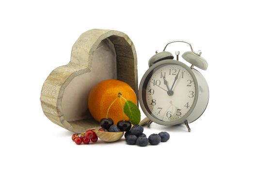 Culinary still life with old-fashioned alarm clock, wooden spoons, cherries and orange, red currants and blueberries over an off white background with copy space
