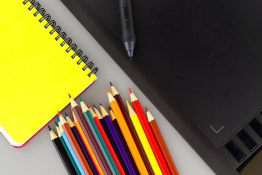 Digital stylus pen on digital graphic tablet for illustrators and designers opposite colored drawing pencils next to a wired notebook . Top view, free space for text
