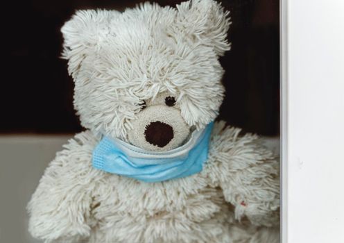 Toy polar bear with medical mask behind window glass. Selective focus