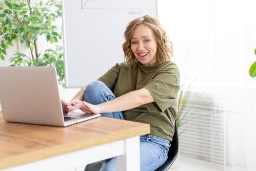Business woman using laptop sitting desk white office interior with houseplant looking camera Business people Business person Online, Young and successful Dresed green shirt blue jeans barefoot relaxing