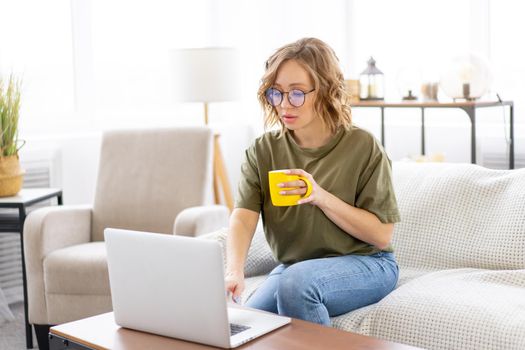 Woman with glasses use laptop typing keyboard sitting couch big window background home interior Freelance female working from home Distance learning student relaxing watch lessons video conference.