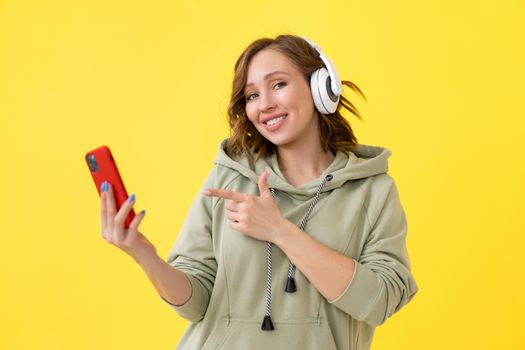 Happy teeth smile woman listen music headphones holding smartphone in hand looking screen Caucasian female enjoy podcast or audio books dressed oversize hoodie yellow background close up portrait