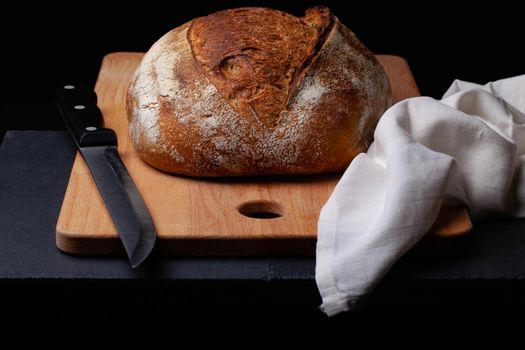 Artisan sourdough bread. Freshly baked round loaf of sourdough bread with linen cloth and knife on black background