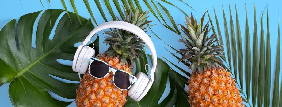 Funny pineapple wearing white headphone, concept of listening music, isolated on blue background with tropical palm leaves, top view, flat lay design.