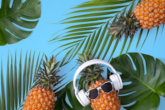 Funny pineapple wearing white headphone, concept of listening music, isolated on blue background with tropical palm leaves, top view, flat lay design.