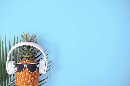 Funny pineapple wearing white headphone, concept of listening music, isolated on blue background with tropical palm leaves, top view, flat lay design.