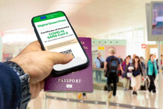 Male hand holding a phone with the European Union vaccination certificate on the screen and a traditional passport inside an airport. Immunity from Covid-19. Travel without restrictions.