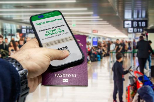 Male hand holding a phone with the European Union vaccination certificate on the screen and a traditional passport inside an airport. Immunity from Covid-19. Travel without restrictions.