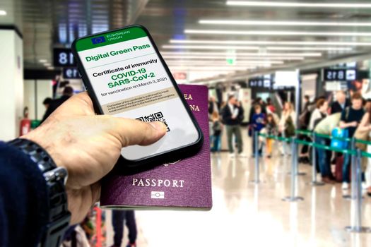 Male hand holding a phone with the European Union vaccination certificate on the screen and a traditional passport inside an airport. Immunity from Covid-19. Travel without restrictions.