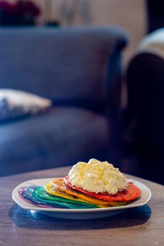 Close-up of a dish with rainbow colored pancakes with a dollop of whipped cream on a wood table in a living room