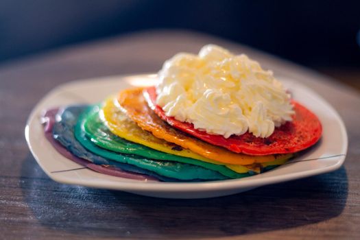 Close-up of a dish with rainbow colored pancakes with a dollop of whipped cream on a wood table