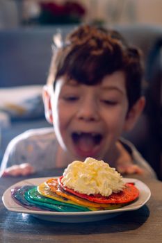 A plate of rainbow colored pancakes  with a dollop of cream on a table with a cute boy looking in the background