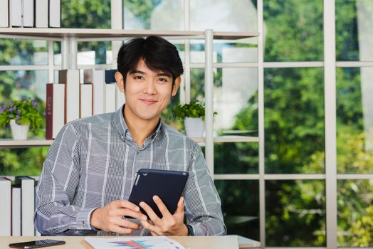 Asian young businessman sitting on desk office he smiles and working on a new tablet computer, the confident young handsome man using touchpad checking his mail