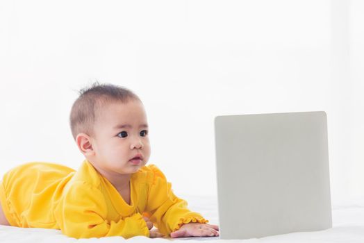 Asian little baby girl wearing a yellow dress typing hands on laptop computer keyboard and looking at monitor on the white bed. Little IT guy