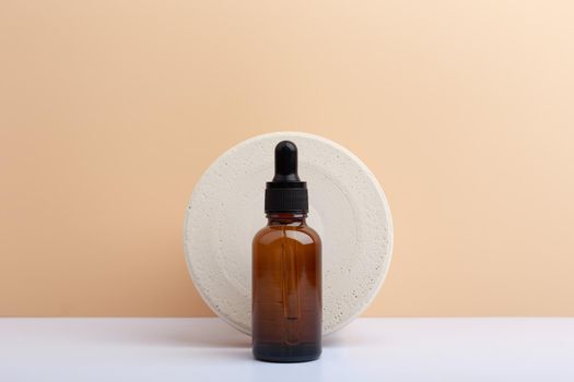 Skin serum in dark brown bottle against round stone circle on white table against beige background with copy space. Concept of hydrating beauty products