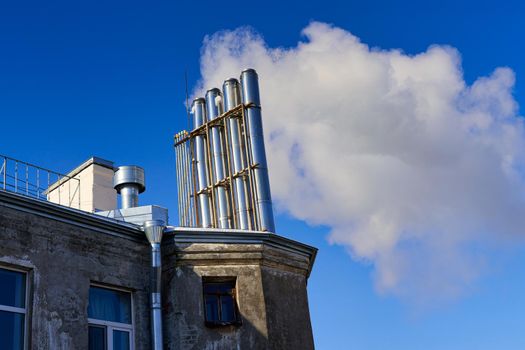 Many chimneys on the roof of an old house smoke against a blue sky. Environmental pollution