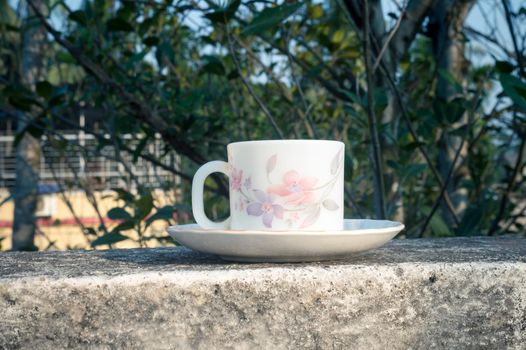 Coffee cup in morning sunlight. Summer fresh cool look. White coffee cup on saucer for hot drink on roof beam of a residential building with bokeh background.