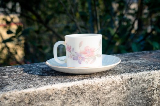 Coffee cup in morning sunlight. Summer fresh cool look. White coffee cup on saucer for hot drink on roof beam of a residential building with bokeh background.