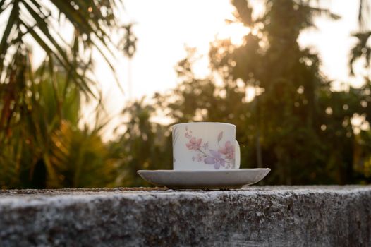 Coffee cup in sunset sunlight. Summer fresh cool look. White coffee cup on saucer for hot drink on roof beam of a residential building with bokeh city in the background.