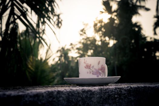 Coffee cup in sunset sunlight. Summer fresh cool look. White coffee cup on saucer for hot drink on roof beam of a residential building with bokeh city in the background.