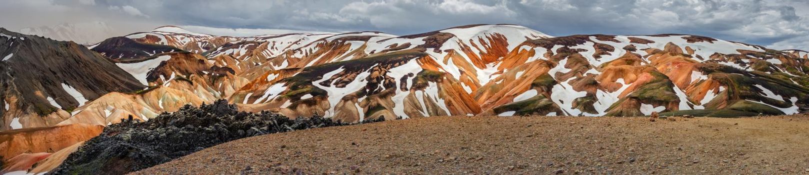 Panoramic amazing Icelandic landscape of colorful rainbow volcanic Landmannalaugar mountains, at famous Laugavegur hiking trail with dramatic snowy sky, and red volcano soil in Iceland.