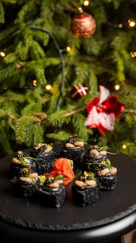 Sushi with black rice, crab meat, avocado, smoked salmon mousse, oar caviar, masago, shrimp cocktail and edible gold leaf with ginger on black table for Christmas with a Christmas tree on background