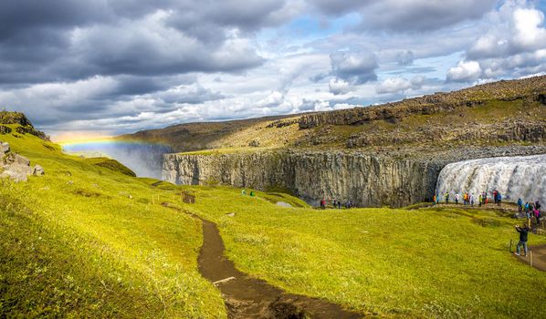 Panoramic view over biggest and most powerful waterfall in Europe called Dettifoss in Iceland, near lake Myvatn, with rainbow at dramatic sky with tourists