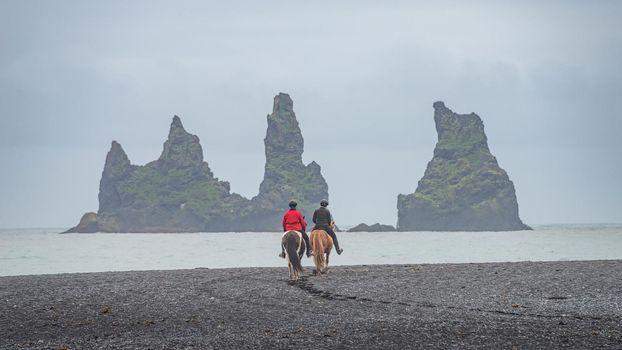 View of basalt stacks Reynisdrangar, black sand beach near Vik and two people in horseback riding, South Iceland