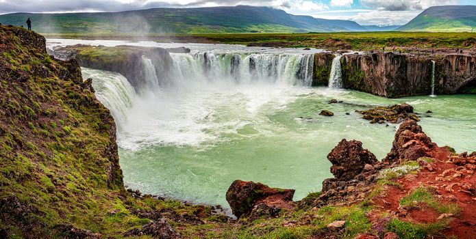 Panoramic view over powerful and wide waterfall Godafoss with a lonely traveler standing at its high cliff, Iceland