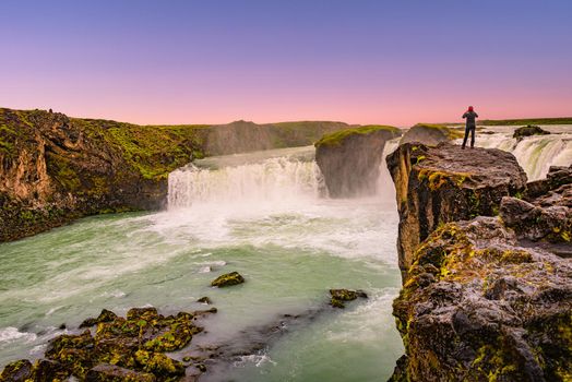 Powerful waterfall Godafoss at beautiful red sunset with a lonely traveler standing at its cliff, Iceland, summer