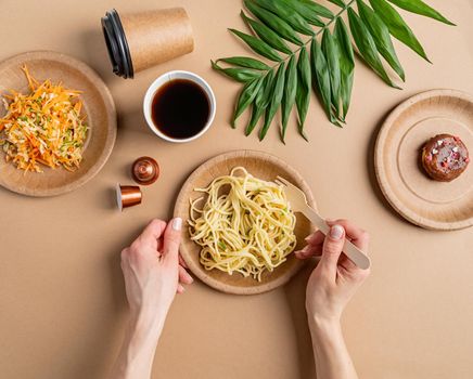 Eco friendly zero waste disposable tableware with pasta, salad and donut top view flat lay on brown background. Hands eating