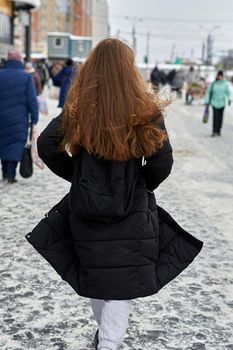 A girl with loose long hair in a black coat tsed on a winter street. Frame from the back