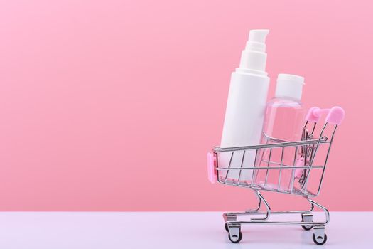 Small toy shopping cart with cosmetic tubes against pink background with copy space. Concept of shopping for beauty products and online sales. Face cream and skin lotion for beauty treatment
