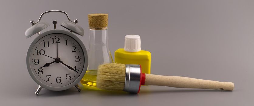 New color, decorating or renovations concept with a still life of an alarm clock with clean paint brush, unlabeled yellow pigment container and laboratory flask over a grey background