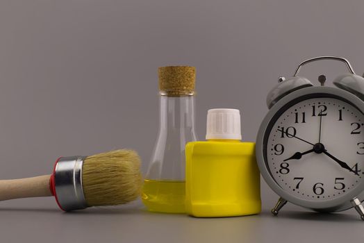 DIY, decorating or renovations concept with a still life of an alarm clock with clean paint brush, yellow pigment container and laboratory flask over a grey background with free space for text
