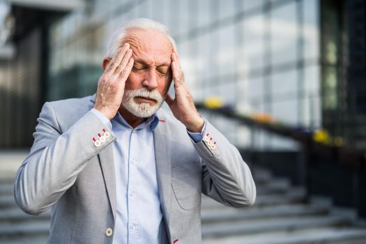 Senior businessman is tired of work. He is standing in front of company building and having headache.
