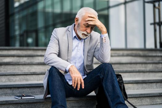 Senior businessman is tired of work. He is sitting in front of company building and having headache.