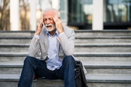 Senior businessman is tired of work. He is sitting in front of company building and having headache.