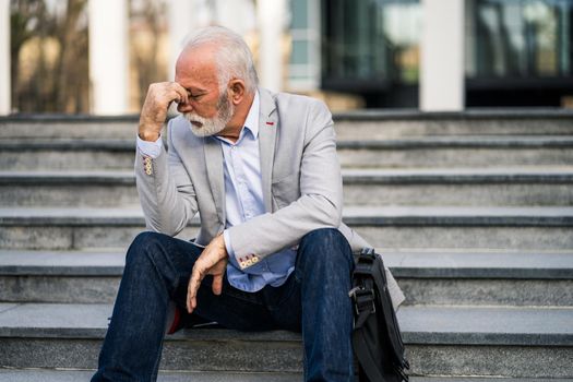 Senior businessman is tired of work. He is sitting in front of company building and having headache.